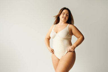 tattooed and brunette curvy woman with plus size body posing in beige bodysuit while standing in studio on grey background, body positive, figure type, self-esteem, tattoo translation: harmony  clipart