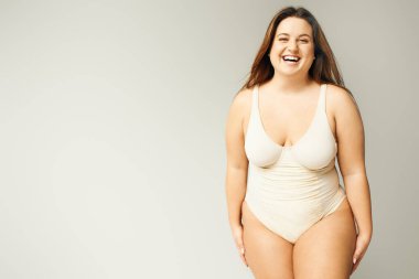 portrait of happy and curvy woman with plus size body posing in beige bodysuit while laughing on grey background, body positive, figure type, looking at camera while standing in studio  clipart