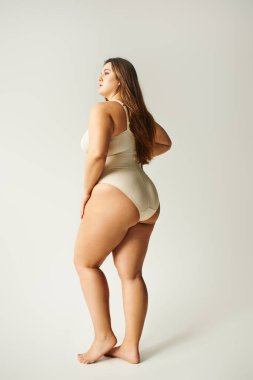 full length of brunette and curvy woman wearing beige bodysuit and standing with bare feet on grey background, body positive, figure type, looking away, body positivity movement  clipart