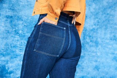cropped view of anonymous plus size woman in orange jacket and posing with hand in pocket of denim jeans while standing on mottled blue background, body positive  clipart