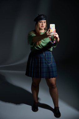 Artı deri bereli, yeşil tişörtlü, ekoseli, zincirli, balıkçı taytı ve siyah ayakkabılı bir kadın. Arka planda akıllı telefondan selfie çekiyor. Dövme çevirisi: