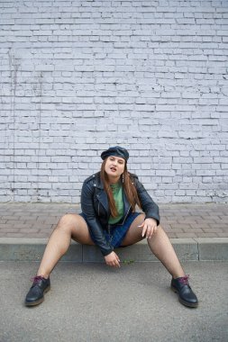 fashionable plus size woman posing in leather jacket and beret, plaid skirt, fishnet tights and black shoes while posing near brick wall on urban street, body positive, full length  clipart