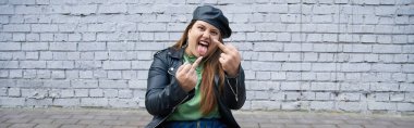 sassy plus size woman sitting in leather jacket and beret while showing middle fingers and sticking tongue out near brick wall on urban street, body positive, bad behavior, banner clipart
