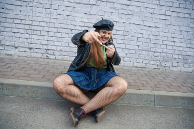 full length of sassy plus size woman posing in leather jacket, beret, plaid skirt with chains, fishnet tights and black shoes while showing middle fingers and smiling near brick wall on urban street  clipart