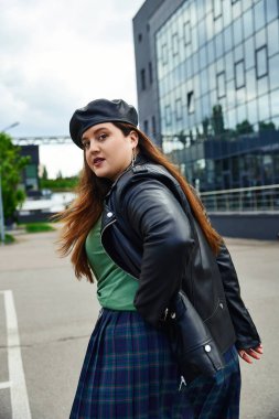 stylish woman with plus size body walking in leather jacket with black beret, plaid skirt and greet t-shirt near blurred modern building on urban street outdoors, body positive, looking at camera clipart