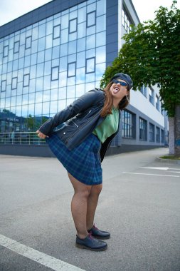 plus size woman in sunglasses, leather jacket with black beret, plaid skirt and fishnet tights sticking out tongue and posing near modern building and tree on urban street, body positive, full length clipart