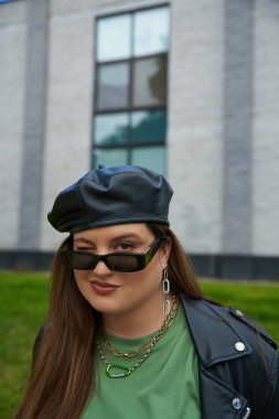 portrait of plus size brunette woman posing in stylish sunglasses, leather jacket with black beret and greet t-shirt winking near blurred modern building on urban street outdoors, body positive  clipart