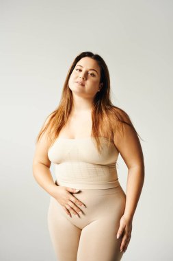 plus size woman with long hair and bare shoulders looking at camera while posing in beige strapless top and underwear in studio isolated on grey background, body positive, self-love  clipart