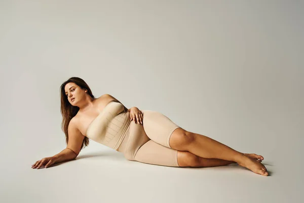 barefoot woman with plus size body in strapless top with bare shoulders and underwear posing while lying in studio on grey background, body positive, self-love, relaxing, looking away