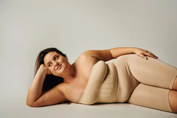 brunette woman with plus size body in strapless top with bare shoulders and underwear posing while lying in studio on grey background, body positive, self-love, relaxing, looking away