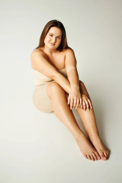full length of barefoot and pretty woman with plus size body in strapless top with bare shoulders and underwear posing while sitting in studio on grey background, body positive, figure type