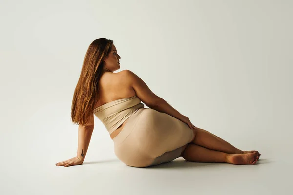 back view of barefoot woman with plus size body in strapless top with bare shoulders and underwear posing while sitting in studio on grey background, body positive, tattoo translation: harmony