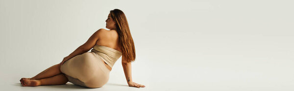 задний вид босиком женщины с плюс размер тела в верхней части без бретелек с обнаженными плечами позирует, сидя в студии на сером фоне, тело положительное, татуировка перевод: гармония, баннер 
