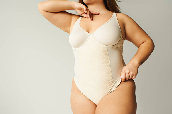 partial view of brunette plus size woman in beige bodysuit posing while standing in studio on grey background, body positive, figure type, self-esteem, tattoo translation: harmony