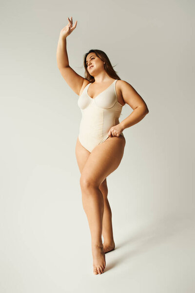 full length of brunette curvy woman with plus size body posing in beige bodysuit while standing with raised hand in studio on grey background, body positive, figure type, smiling while looking away 