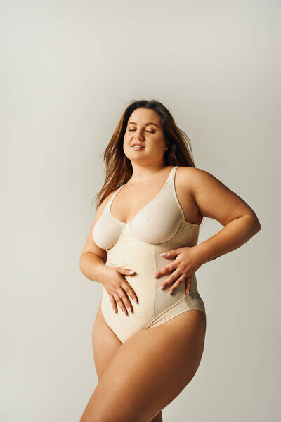 brunette and curvy woman with tattoo wearing beige bodysuit and standing with hands on waist on grey background, body positive, figure type, body positivity movement, closed eyes 