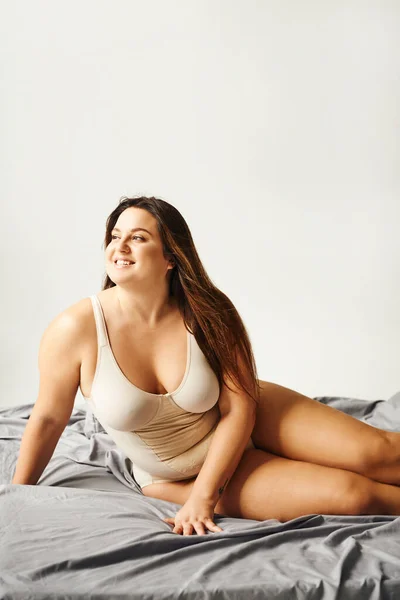 charming woman with plus size body wearing beige bodysuit and smiling and posing on bed with grey bedding, body positive, figure type, bare feet, looking away, tattoo translation: harmony