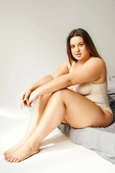 full length of charming woman with plus size body and bare feet wearing beige bodysuit and looking at camera while sitting on bed with grey bedding, body positive, figure type