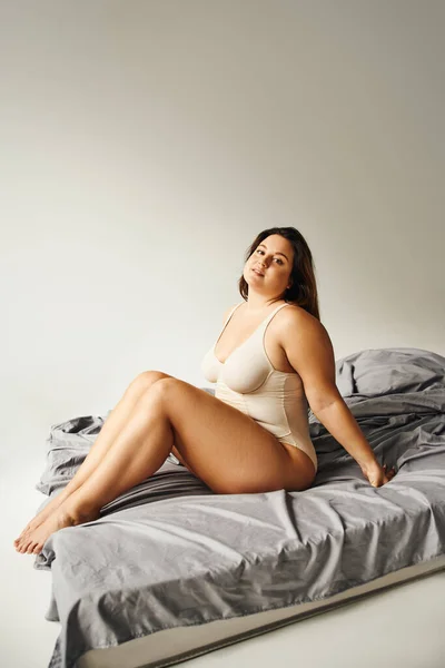 full length of alluring woman with long hair and plus size body and bare feet wearing beige bodysuit and looking at camera while sitting on bed with grey bedding, body positive, figure type