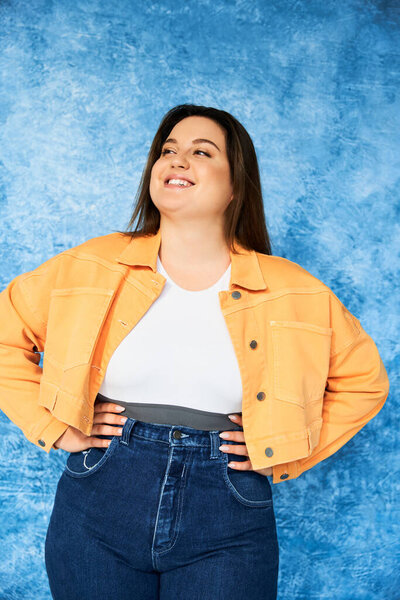 cheerful plus size woman with long hair and natural makeup wearing crop top, orange jacket and denim jeans while posing with hands on hips and looking away on mottled blue background, body positive 