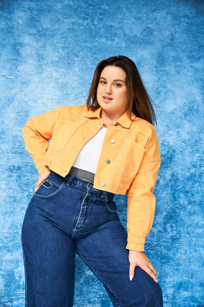 woman with plus size body and long hair, wearing crop top, orange jacket and denim jeans while posing with hand on hip and looking at camera on mottled blue background, body positive 