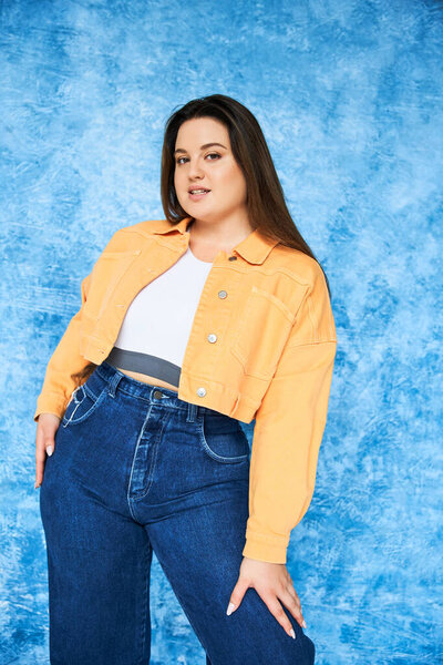 plus size woman with brunette long hair and natural makeup wearing crop top, orange jacket and denim jeans while posing and looking at camera on mottled blue background, body positive 