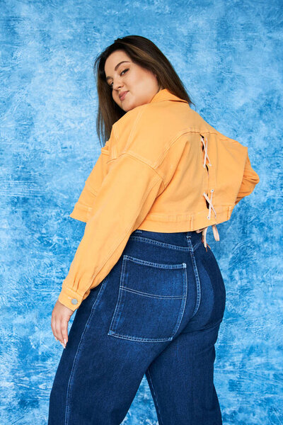 alluring plus size woman with long hair and natural makeup wearing crop top, orange jacket and denim jeans while posing and looking at camera on mottled blue background, body positive 