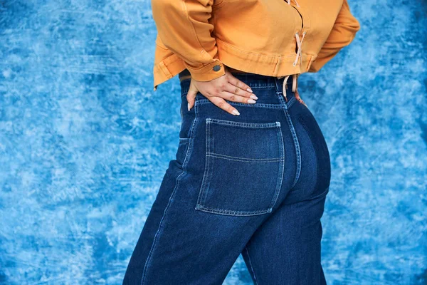 stock image cropped view of anonymous plus size woman in orange jacket and denim jeans posing with hand on hip while standing with hand on hip on mottled blue background, body positive 