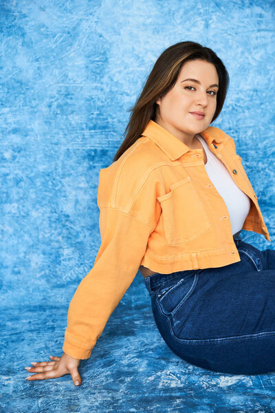 body positive and pretty plus size woman with long hair and natural makeup wearing crop top, orange jacket and denim jeans while sitting and looking at camera on mottled blue background 