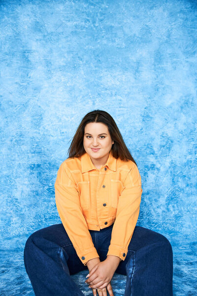 body positive, plus size woman with brunette hair and natural makeup sitting in orange jacket and denim jeans while smiling and looking at camera on mottled blue background 