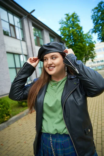 一个快乐的女人 身穿皮夹克 穿着黑色贝雷帽 紧靠着模糊的现代建筑和户外街道上的树木 身强体壮 — 图库照片