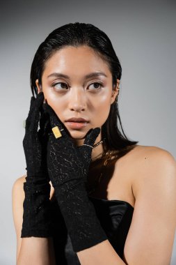portrait of attractive and asian young woman with short hair posing in black strapless dress and gloves with golden rings, looking away on grey background, wet hairstyle, necklaces, natural makeup clipart