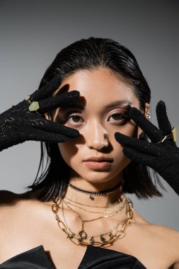 portrait of brunette and asian young woman with short hair posing in black gloves with golden rings, looking at camera on grey background, wet hairstyle, hands near face, natural makeup clipart