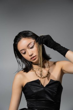 portrait of brunette and asian young woman with short hair posing in black gloves and strapless dress while holding golden jewelry on grey background, wet hairstyle, natural makeup clipart