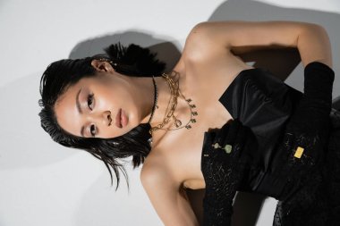 top view of charming asian young woman with short hair lying in black gloves and strapless dress while posing in golden jewelry on grey background, wet hairstyle, natural makeup, looking at camera clipart