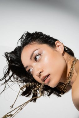 Göz kamaştırıcı Asyalı model, kısa esmer saçlı, altın mücevherleri ağzında tutarken gri arka planda yatıyor, günlük makyaj, ıslak saç stili, genç kadın, başka tarafa bakıyor.