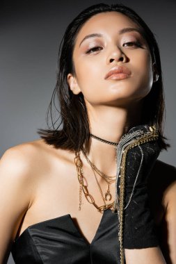 portrait of brunette and asian young woman with short hair holding golden and silver jewelry while wearing glove and standing in black strapless dress grey background, wet hairstyle, natural makeup clipart