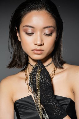 portrait of alluring asian young woman with short hair holding golden and silver jewelry while wearing glove and standing in strapless dress grey background, wet hairstyle, natural makeup clipart