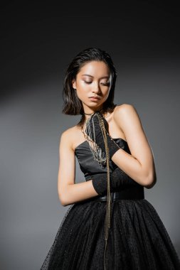 charming asian young woman with short hair holding golden and silver jewelry while wearing gloves and standing in elegant strapless dress on grey background, wet hairstyle, natural makeup clipart