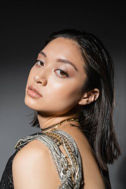 portrait of brunette and asian young woman with short hair posing with golden and silver jewelry on shoulder and looking at camera on grey background, wet hairstyle, natural makeup clipart