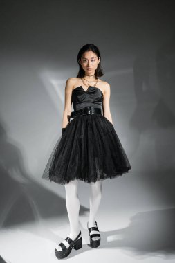 full length of stylish asian young woman with short hair posing in black strapless dress, white tights and shoes while looking at camera on grey background, wet hairstyle, golden necklaces  clipart