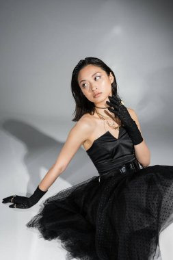 chic asian young woman with short hair sitting in black strapless dress with tulle skirt with belt and gloves while looking away and touching neck on grey background, wet hairstyle, golden necklaces  clipart