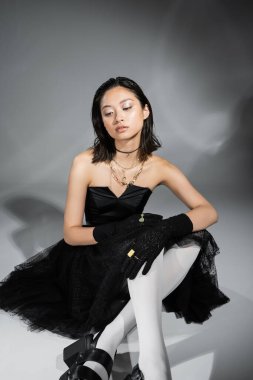 dreamy asian young woman with short hair sitting in black strapless dress with tulle skirt white tights, shoes and gloves while looking away on grey background, wet hairstyle, golden necklaces  clipart