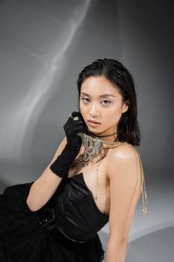 asian model with short and brunette hair holding golden jewelry in hand while posing in strapless dress and black glove on grey background, everyday makeup, wet hairstyle, young woman, glamour  clipart