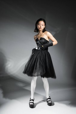 full length of stylish asian young woman with short hair standing in black strapless dress with tulle skirt and belt, gloves, white tight and shoes on grey background, wet hairstyle, golden necklaces  clipart