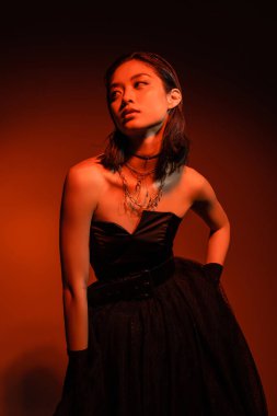 asian woman with short hair and wet hairstyle posing with hand on hip in black strapless dress with tulle skirt and gloves while standing on orange background with red lighting, golden jewelry  clipart