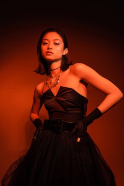 dreamy asian woman with short hair and wet hairstyle posing in black strapless dress with tulle skirt and gloves while standing on orange background with red lighting, golden jewelry, young model clipart