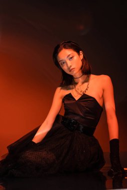 beautiful asian woman with short hair and wet hairstyle posing in strapless dress with tulle skirt and gloves while standing on orange background with red lighting, young model, looking at camera  clipart