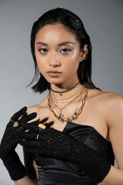 stock image portrait of brunette and asian young woman with short hair posing in black strapless dress and gloves with golden rings, looking at camera on grey background, wet hairstyle, necklaces, natural makeup
