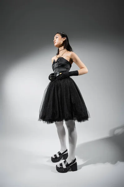 stock image full length of alluring and asian young woman with short hair posing in black strapless dress with tulle skirt and gloves, looking away on grey background, wet hairstyle, golden necklaces 